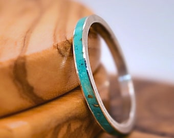 Könnte beinhalten: Ein silberner Ring mit Türkiseinlage liegt auf einer Holzoberfläche. Das Band des Rings ist dünn, und der Türkis hat ein gesprenkeltes Aussehen. Die Holzmaserung ist sichtbar, mit warmen Brauntönen.