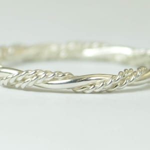 Puede incluir: Un anillo plateado con un diseño de cuerda trenzada. El anillo está hecho de un material metálico brillante y tiene una forma circular continua. El patrón trenzado crea una superficie texturizada, agregando interés visual a la joya.