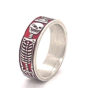 May include: Silver ring with a red band featuring a white skeleton design. The ring has a skull and rib cage motif, with intricate detailing. The ring is set against a white background.