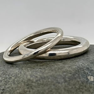 May include: Two silver rings, one stacked atop the other, are displayed on a gray stone. The rings have a smooth, polished finish, reflecting the light. The rings are a simple, elegant design, suitable for everyday wear.