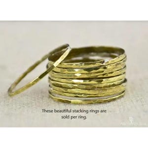 May include: A stack of gold-colored stacking rings with a hammered texture. One ring is separated from the stack, resting on a neutral surface. The rings are a simple, minimalist design. Text on the image reads: "These beautiful stacking rings are sold per ring."