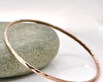 May include: A hammered copper bangle bracelet. The bracelet is a thin, circular band with a textured surface. The copper has a warm, reddish-brown color. The bracelet is resting on a gray stone.