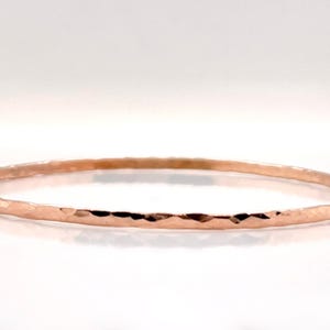 May include: A rose gold bangle bracelet with a hammered texture. The thin, circular bracelet is displayed on a white background. The jewelry piece has a simple, elegant design.