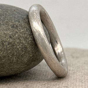 May include: A silver ring with a textured surface. The ring is round and appears to be made of metal. The ring is resting on a gray stone. The ring is a simple, elegant piece of jewelry.
