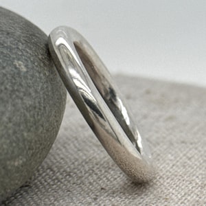 May include: A polished silver ring with a rounded rectangular shape. The ring is resting on a gray stone and a light gray fabric background. The ring's surface reflects light, highlighting its smooth texture.