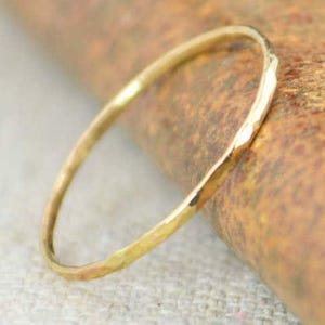Könnte beinhalten: Ein zarter, gehammerter Goldring. Das dünne Band hat eine strukturierte Oberfläche und ein schlichtes, elegantes Design. Der Ring ist vor einem verschwommenen, neutralen Hintergrund platziert.