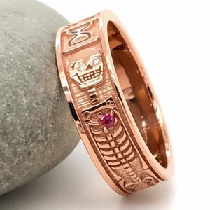 Op de afbeelding: Roségouden ring met een gedetailleerd skeletontwerp. De ring heeft een schedel met diamantaccenten en een kleine rode edelsteen. De band is gegraveerd met een ribbenkast en andere decoratieve elementen. De ring wordt getoond op een grijze steen.