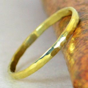 May include: A hammered gold ring with a textured surface. The ring is a thin band, and the gold has a warm, yellow tone. The ring is resting on a textured, orange and brown surface.