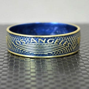 Puede incluir: Un anillo azul y dorado con el texto "LOS ANGELES" alrededor de la banda. El anillo tiene un patrón texturizado y un borde dorado. El interior del anillo es de color azul oscuro.