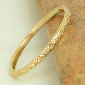Puede incluir: Un anillo dorado con un delicado diseño floral. El anillo es delgado y descansa sobre una superficie marrón texturizada. El diseño del anillo presenta pequeños motivos florales y de hojas repetidos.