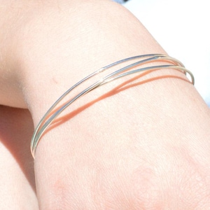 May include: A silver bangle bracelet with three thin bands is worn on a wrist. The bracelet is the focal point, with the person's arm and clothing in the background. The person is wearing a light blue button-down shirt and blue jeans.