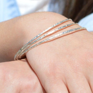 May include: A close-up of three silver bangles with a textured surface. The bangles are stacked on a wrist, with another hand resting below. The person is wearing a light blue shirt and jeans.