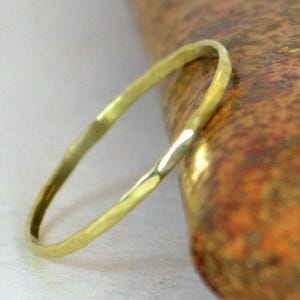May include: A delicate, hammered gold ring. The thin band has a textured surface and a warm, yellow hue. The ring is set against a neutral background and a textured, reddish-brown surface.