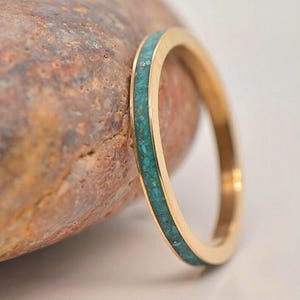 Op de afbeelding: Een goudkleurige ring met een band van turquoise gekleurde stenen. De ring is geplaatst tegen een getextureerde, bruine steen en een wit oppervlak. Het ontwerp is eenvoudig en elegant, waarbij het contrast tussen goud en turquoise wordt benadrukt.