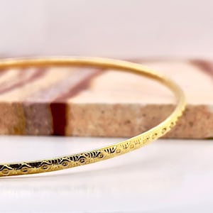 May include: A gold-colored bangle bracelet with an intricate, repeating pattern. The bracelet is thin and circular, with a detailed design along its surface. The background features a textured, multi-colored surface.