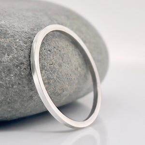 Puede incluir: Un anillo minimalista de plata con una banda plana. El anillo está colocado sobre una piedra gris, con un fondo blanco. El diseño sencillo del anillo lo hace adecuado para el uso diario.