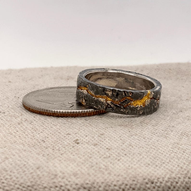 Puede incluir: Un anillo de plata con un exterior texturizado gris oscuro y una incrustaci&oacute;n dorada. El anillo est&aacute; junto a una moneda, mostrando su tama&ntilde;o. El dise&ntilde;o presenta una est&eacute;tica org&aacute;nica y robusta. Di&aacute;metro: aprox. 2 cm.