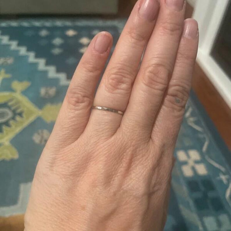 Op de afbeelding: Een dunne, zilverkleurige ring wordt op een vinger gedragen. De ring heeft een eenvoudig, minimalistisch ontwerp. De hand staat centraal, met de ring als hoofdonderwerp. De achtergrond is wazig, met patronen.