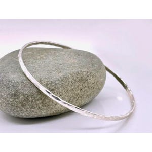 May include: A silver bangle bracelet with a hammered texture rests on a gray stone. The bracelet is circular and appears to be made of metal. The stone is smooth and oval-shaped. The background is a soft white.
