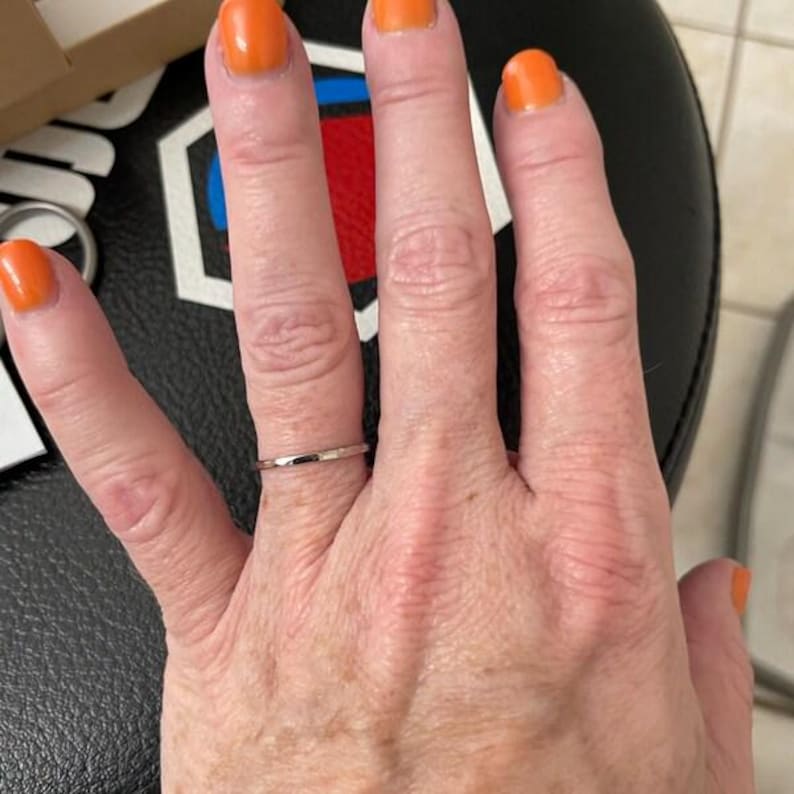 Op de afbeelding: Een dunne zilveren ring wordt aan een vinger gedragen. De ring heeft een eenvoudig ontwerp. De nagels van de persoon zijn oranje gelakt. De achtergrond is een zwart oppervlak met een wit en rood ontwerp.