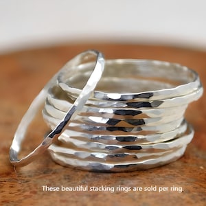 May include: A stack of hammered silver stacking rings. One ring is leaning against the stack. The rings have a textured, faceted surface. The rings are displayed on a brown surface.