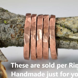 May include: Close-up of six copper rings with a hammered texture, displayed on a weathered tree branch. The rings are a warm, reddish-brown color. The text at the bottom says "These are sold per Ring, Handmade just for you!"