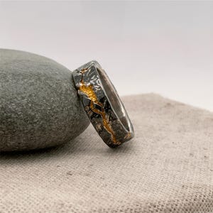 Puede incluir: Un anillo de plata con un exterior texturizado gris oscuro y un dise&ntilde;o dorado en forma de grieta. El anillo es rectangular y descansa sobre una piedra gris y una tela beige.