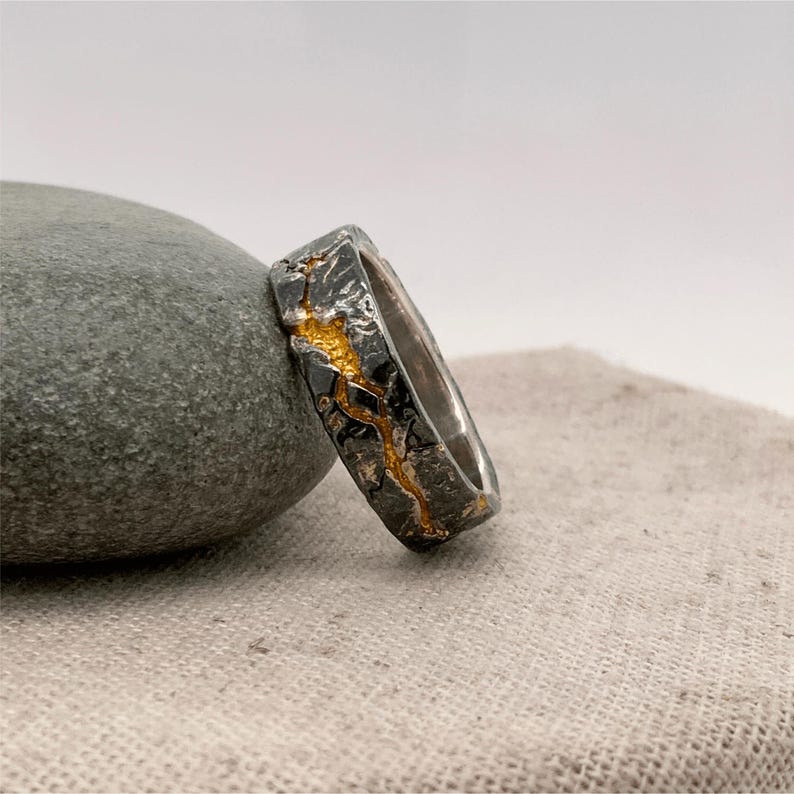 Puede incluir: Un anillo de plata con un exterior texturizado gris oscuro y un dise&ntilde;o dorado en forma de grieta. El anillo es rectangular y descansa sobre una piedra gris y una tela beige.