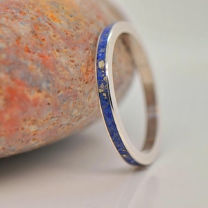 Op de afbeelding: Een zilveren ring met een band ingelegd met blauwe en goudkleurige lapis lazuli. De ring is geplaatst tegen een getextureerde, veelkleurige steen. Het ontwerp van de ring is eenvoudig en elegant, geschikt voor dagelijks gebruik.
