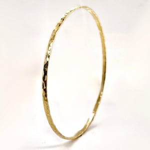 以下が含まれることがあります： ハンマー加工されたゴールドのバングルブレスレット。細い円形のブレスレットは、わずかに不規則な形状のテクスチャ加工された表面を持っています。ゴールドの色は明るく光沢があり、光を反射します。ジュエリーは白い背景に配置されています。