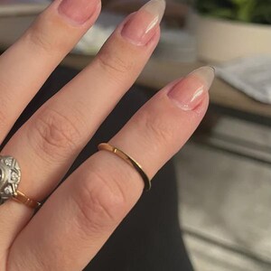 May include: A close-up of a hand with long, natural nails and a thin, gold band ring on the ring finger. Another ring with a decorative setting is visible on the adjacent finger. The nails are a light pink color.