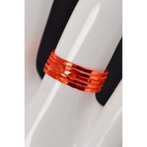 May include: A vibrant orange ring composed of multiple bands, creating a textured appearance. The ring is displayed on a white hand, with a black background. The ring's design is simple yet eye-catching.