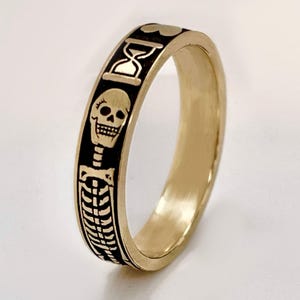 Op de afbeelding: Een goudkleurige ring met zwarte geëtste ontwerpen. De ring heeft een herhalend patroon van een schedel, zandloper en ribbenkast. De ring is waarschijnlijk gemaakt van metaal en heeft een gladde, gepolijste afwerking.