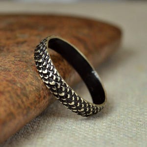 Può includere: Un anello strutturato con interno nero e un motivo di piccole forme bianche a forma di scaglia all'esterno. L'anello è posto su uno sfondo neutro sfocato, evidenziando il suo design e la sua maestria unici.