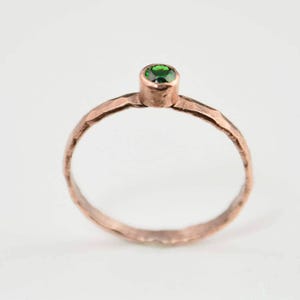 Puede incluir: Un anillo de color cobre con textura martillada. El anillo presenta una pequeña gema verde redonda y brillante engastada en un bisel elevado. La banda tiene un diseño simple y elegante.