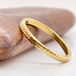 May include: A gold ring with a textured, leaf-like pattern. The ring is set against a blurred, brown and white background. The ring's design is visible, showcasing its intricate details.