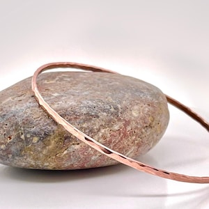 May include: A hammered copper bangle bracelet rests on a textured, oval-shaped stone. The bracelet has a simple, elegant design with a slightly irregular surface. The stone is a mix of brown, gray, and pink hues, creating a natural contrast.