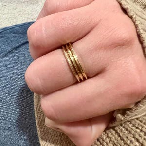May include: A close-up view of three thin, gold-colored rings stacked on a finger. The rings have a simple, minimalist design and appear to be made of metal. The rings are worn on a finger, with the hand and part of a sleeve visible.