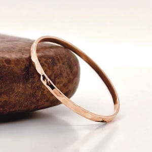 Könnte beinhalten: Ein gehämmertes Armreif-Armband aus Roségold. Das Armband hat ein einfaches, kreisförmiges Design mit strukturierter Oberfläche. Der Armreif liegt auf einem braunen, strukturierten Stein mit weißem Hintergrund.