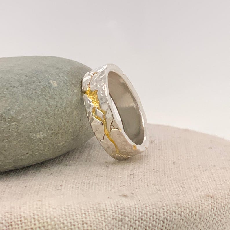 Puede incluir: Un anillo de plata con una superficie texturizada y detalles dorados. El anillo tiene un dise&ntilde;o &uacute;nico con una apariencia agrietada, y el oro rellena las grietas. El anillo se muestra sobre una superficie de color neutro.