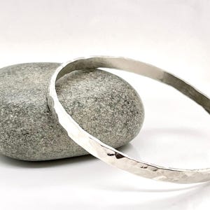 May include: A hammered silver bangle bracelet. The bracelet has a textured surface and a simple, elegant design. The bracelet is displayed on a gray stone against a white background. This piece is suitable for everyday wear.