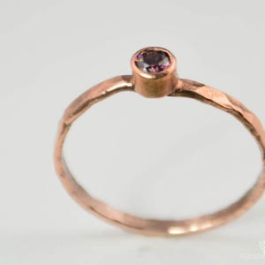 Puede incluir: Un anillo de cobre martillado con una pequeña gema redonda y púrpura. El anillo tiene una banda texturizada y un diseño simple y elegante. La gema está engastada en un bisel.