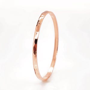 Könnte beinhalten: Ein gehämmertes Armreif-Armband aus Roségold. Das dünne, kreisförmige Armband hat eine strukturierte Oberfläche mit reflektierenden Facetten. Die warme, metallische Farbe des Armbands kontrastiert mit dem weißen Hintergrund. Ein Schmuckstück.