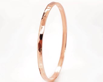 May include: A hammered rose gold bangle. The thin, circular bracelet has a textured surface with reflective facets. The warm, metallic colour of the bracelet contrasts against the white background. The bangle is a piece of jewellery.