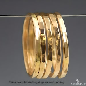 May include: A set of five gold-colored stacking rings. Each ring has a hammered texture, creating a unique, reflective surface. The rings are displayed against a neutral gray background. Text at the bottom reads: "These beautiful stacking rings are sold per ring."
