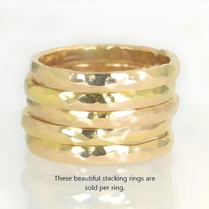 May include: A stack of five gold-colored stacking rings. Each ring has a hammered texture, creating a unique, reflective surface. The rings are displayed on a white background. Text at the bottom reads: "These beautiful stacking rings are sold per ring."