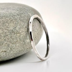 May include: A silver ring with a hammered texture sits next to a gray stone. The ring is thin and has a slightly irregular shape. The background is white.