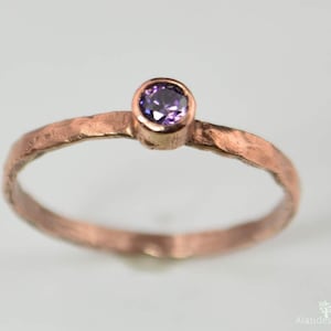 Könnte beinhalten: Ein kupferfarbener Ring mit gehämmerten Texturen und einem runden, lila Edelstein. Der Ring hat ein schlichtes Band und eine Fassung für den Edelstein. Der Ring ist ein Schmuckstück.