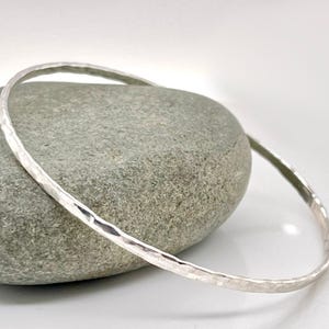 May include: A silver bangle bracelet with a hammered texture rests on a grey stone. The bracelet has a simple, elegant design, reflecting light. The background is a soft white, highlighting the jewellery.