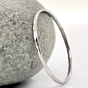 Op de afbeelding: Een zilveren armband met een gehamerde textuur ligt tegen een grijze steen. De armband is een eenvoudig, elegant ontwerp, perfect voor dagelijks gebruik. De armband is gemaakt van een glanzend, reflecterend metaal.